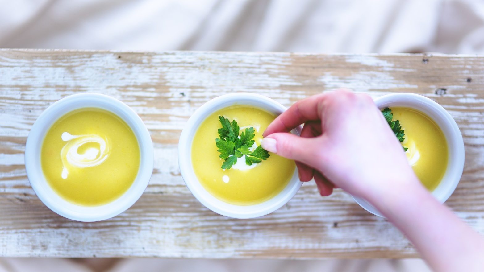 3 pratos de sopas amarelas uma ao lado da outra e uma mão feminina colocando uma folha de salsinha na sopa do meio.