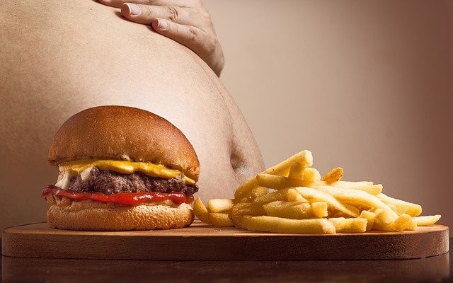 Uma pessoa com obesidade atrás de uma mesa que tem um hambúrguer e uma porção de batata frita.